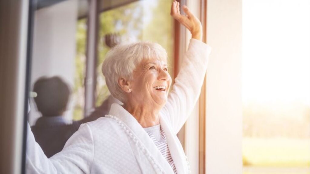 Aged Woman Standing By The Window Smiling